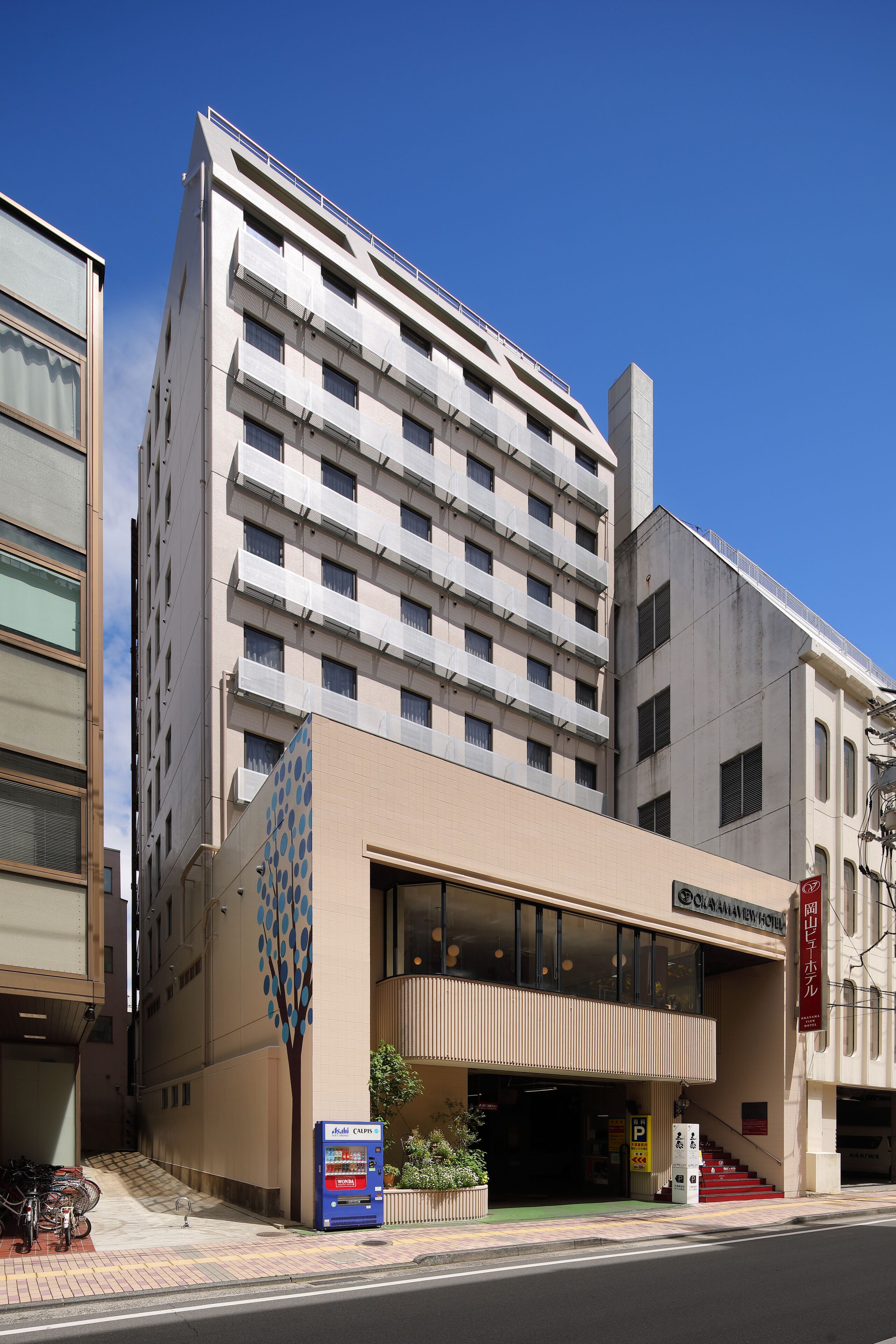 Ein mehrstöckiges Hotel mit moderner Fassade, einer überdachten Eingangsparte und einem Parkplatz mit Verkaufsautomaten.