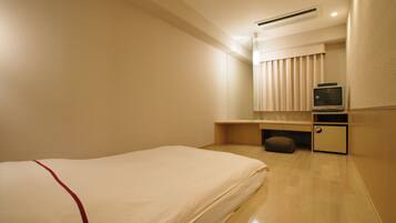 A hotel room with a single bed, a small desk with a television, and a window with blinds.
