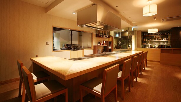 A modern kitchen with a central island, wooden chairs, and built-in shelves with bottles.