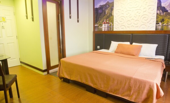 Una habitación de hotel con una cama, una silla, suelo de madera, una puerta y una pared con un panel decorativo y un cuadro enmarcado.