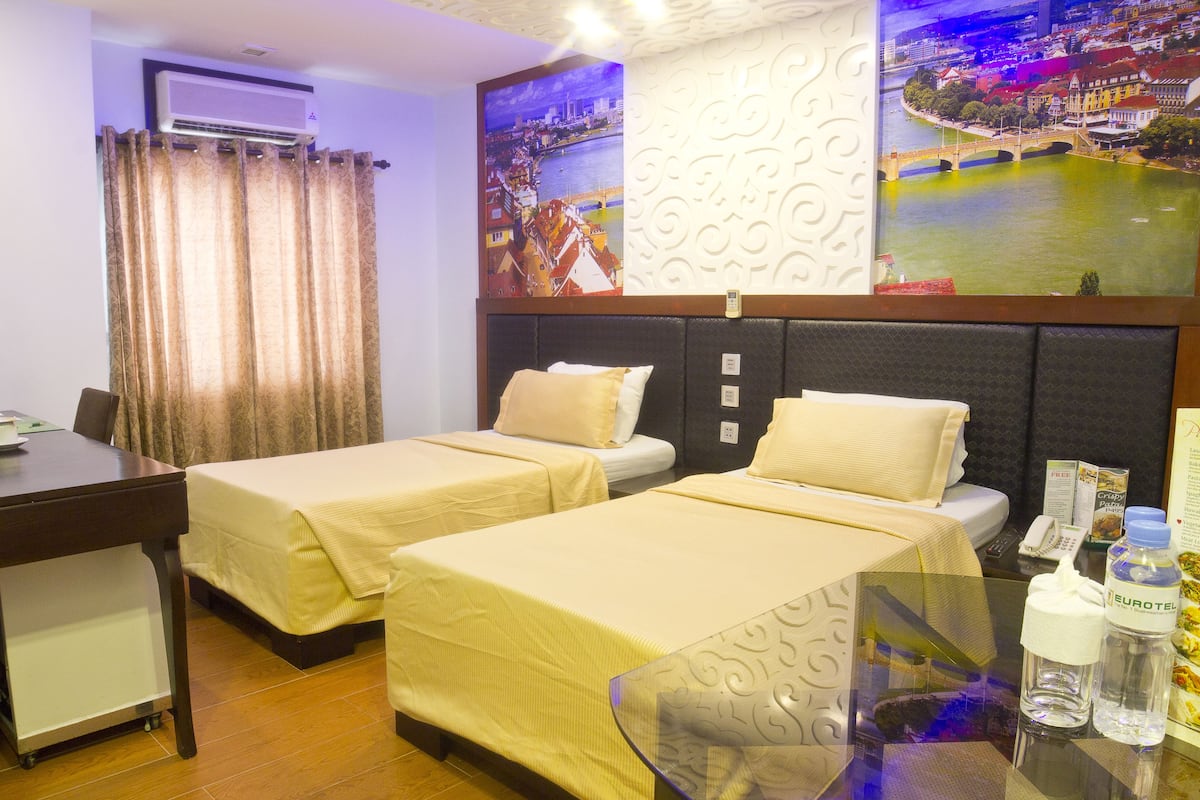 A hotel room with two beds, a desk, and a view of a cityscape.