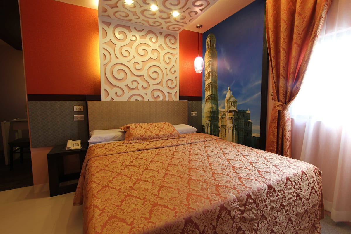 A hotel room with a large bed, a headboard, a wall with a decorative pattern, a painting of a famous tower, and a window with curtains.