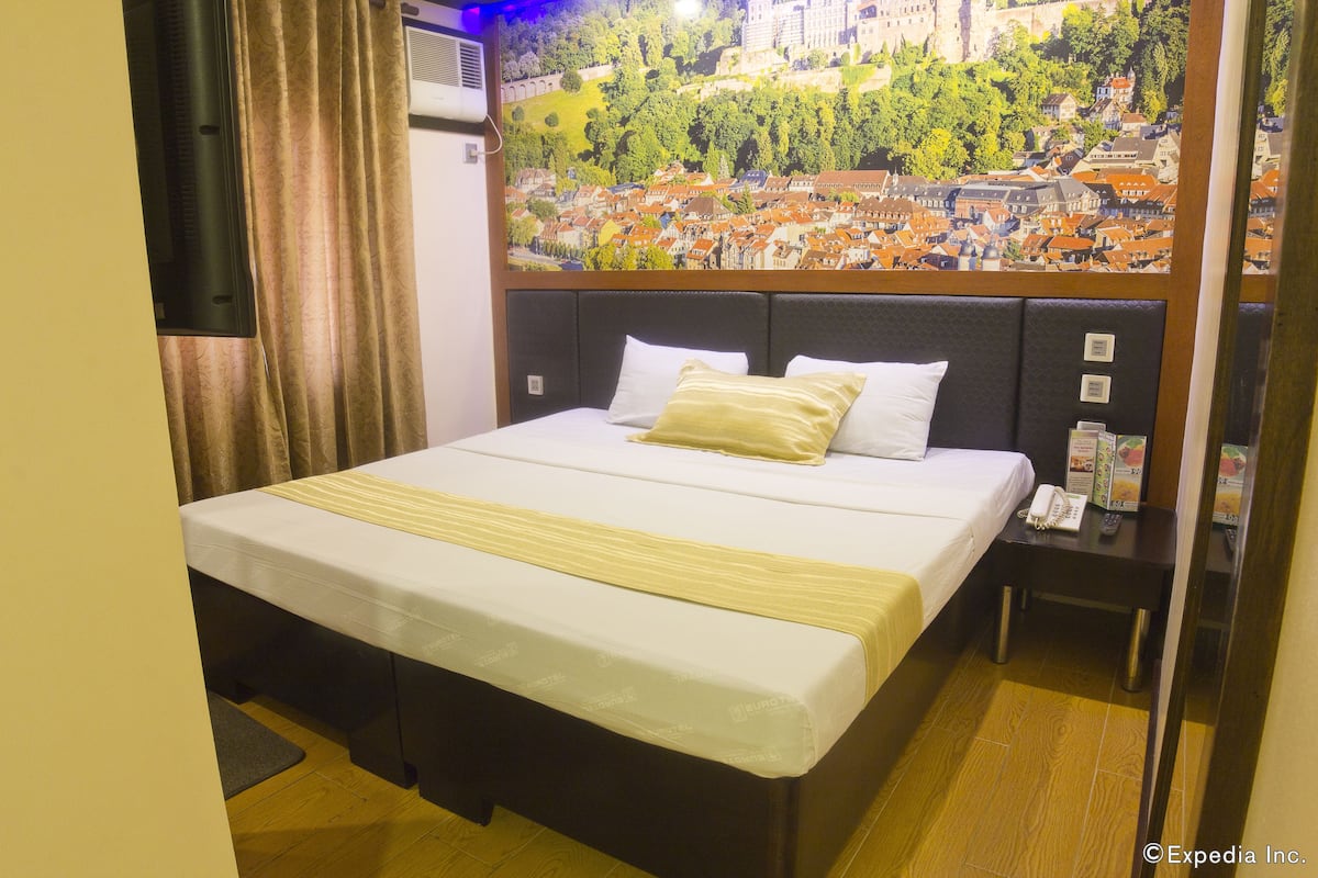 A hotel room with a bed, a nightstand, a telephone, and a wall mural of a cityscape.