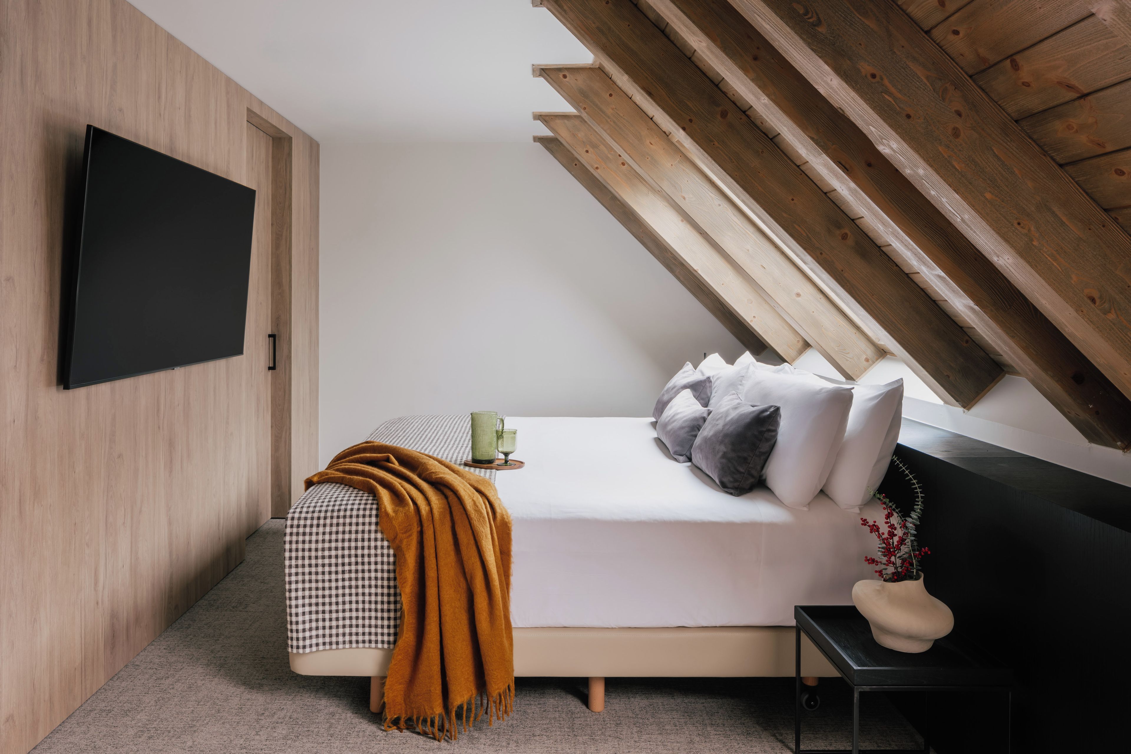 Ein modernes Schlafzimmer mit schrägem Dach, einem flachbildfernseher an der Wand, einem Bett mit kariertem Überwurf, Kissen und einem Nachttisch mit Tasse und Blumenvase.