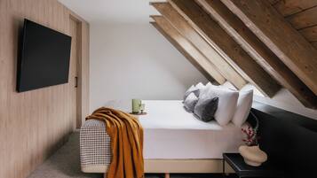 Ein modernes Schlafzimmer mit schrägem Dach, einem flachbildfernseher an der Wand, einem Bett mit kariertem Überwurf, Kissen und einem Nachttisch mit Tasse und Blumenvase.