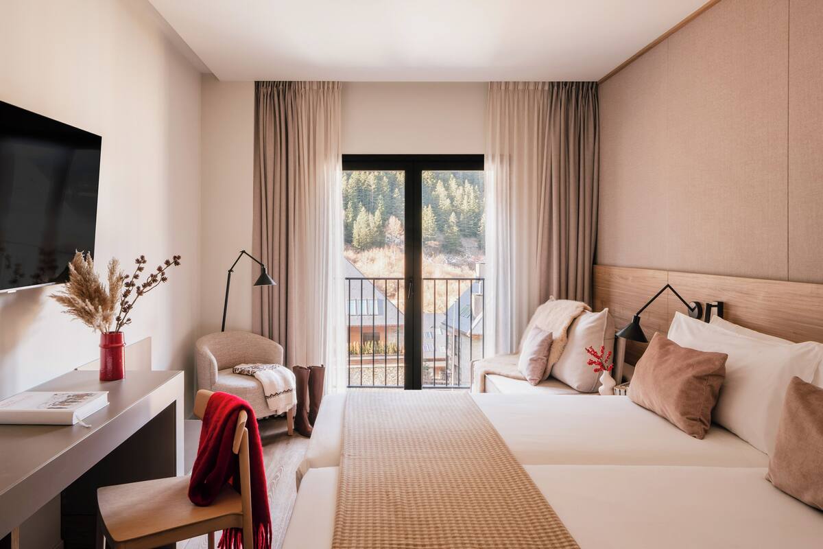 A hotel room with a bed, a desk, a chair, a TV, and a view of trees and buildings outside.