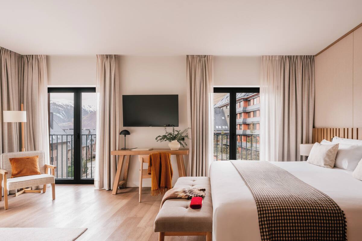A modern hotel room with a large bed, a desk with a chair, a TV, and a view of buildings and mountains.