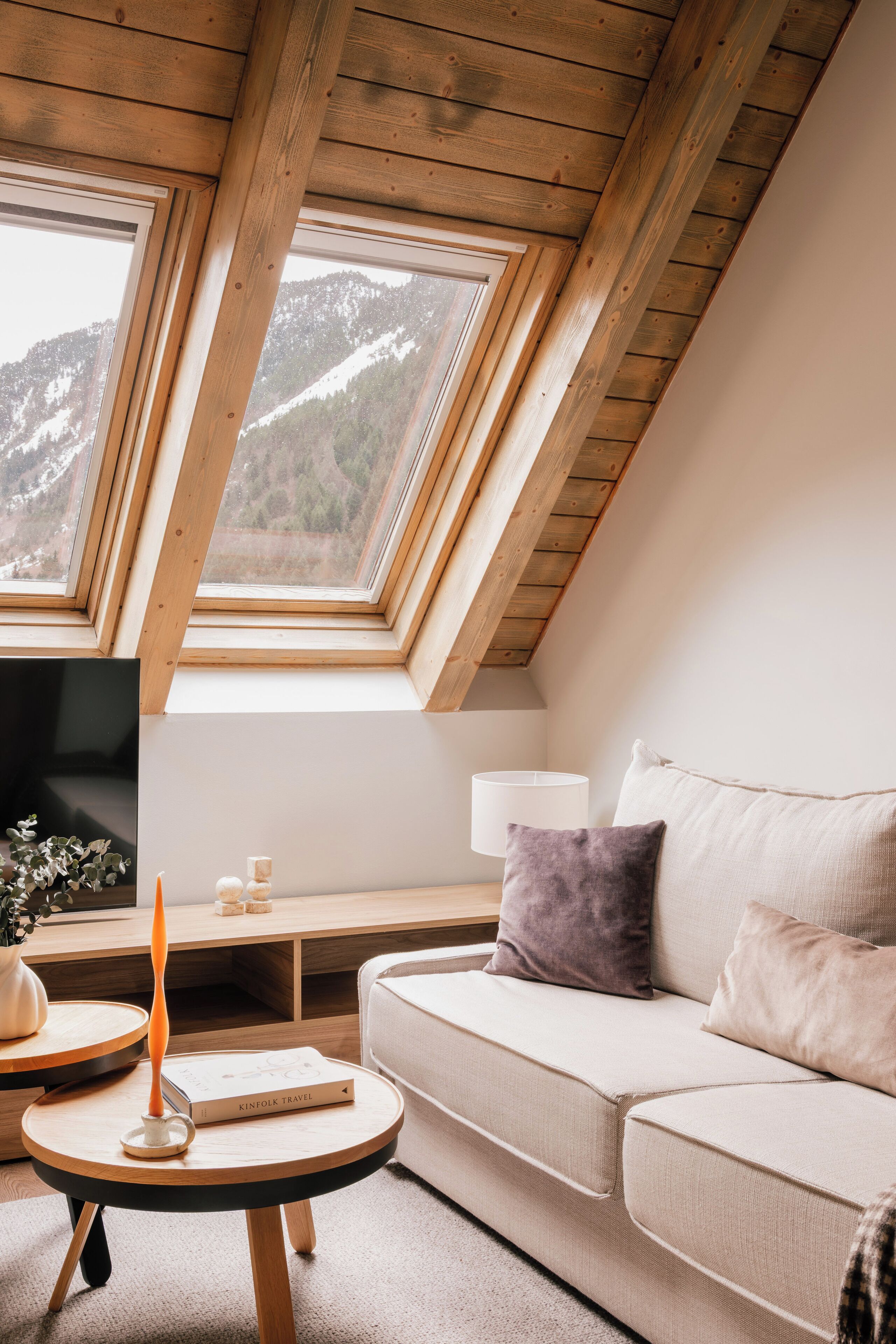 Een knus loft slaapkamer met een schuine plafond, een bank, een klein tafeltje met een kaars, en uitzicht op bergen door dakramen.