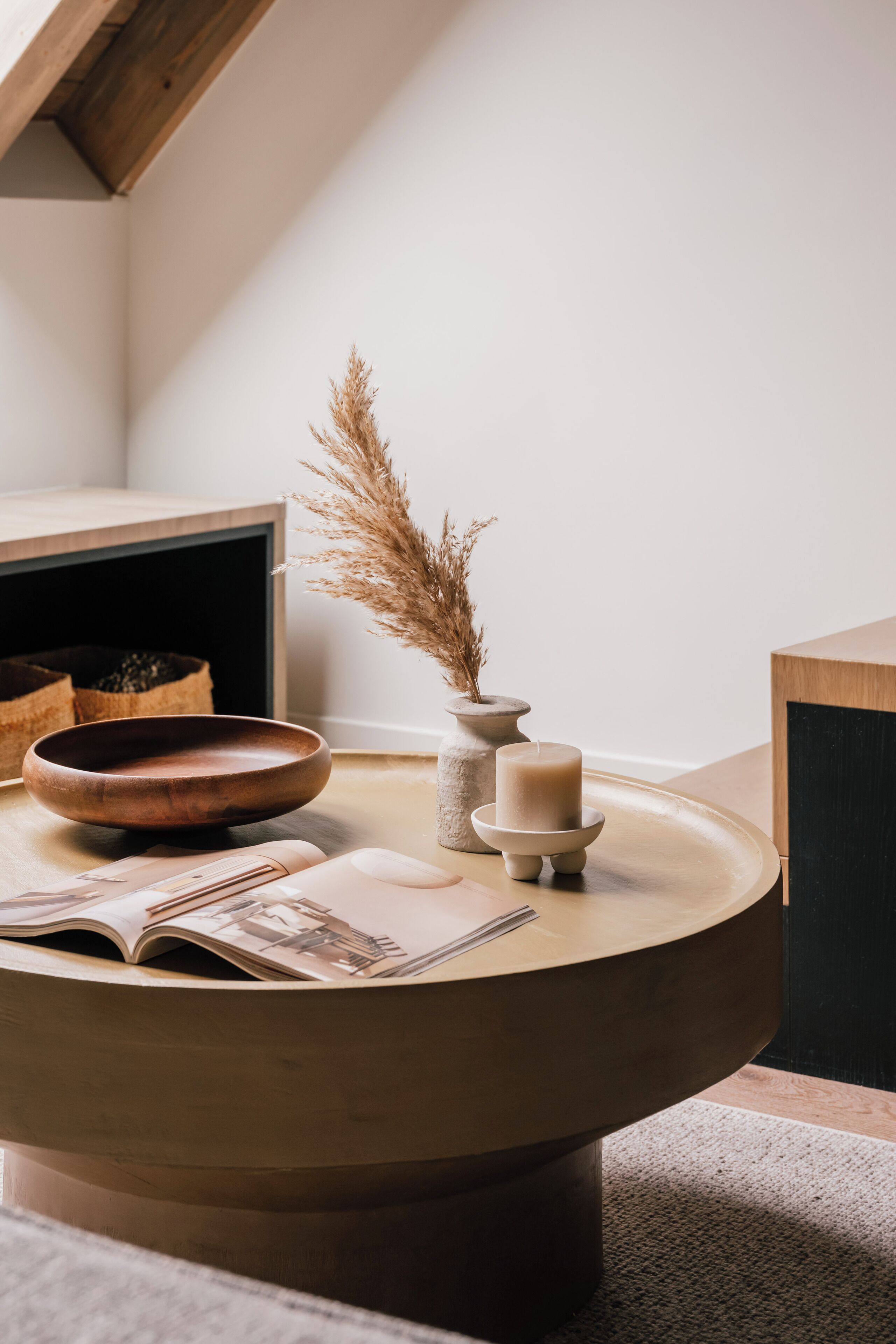 A round wooden table with a bowl, a candle, and a vase with dried plants.
