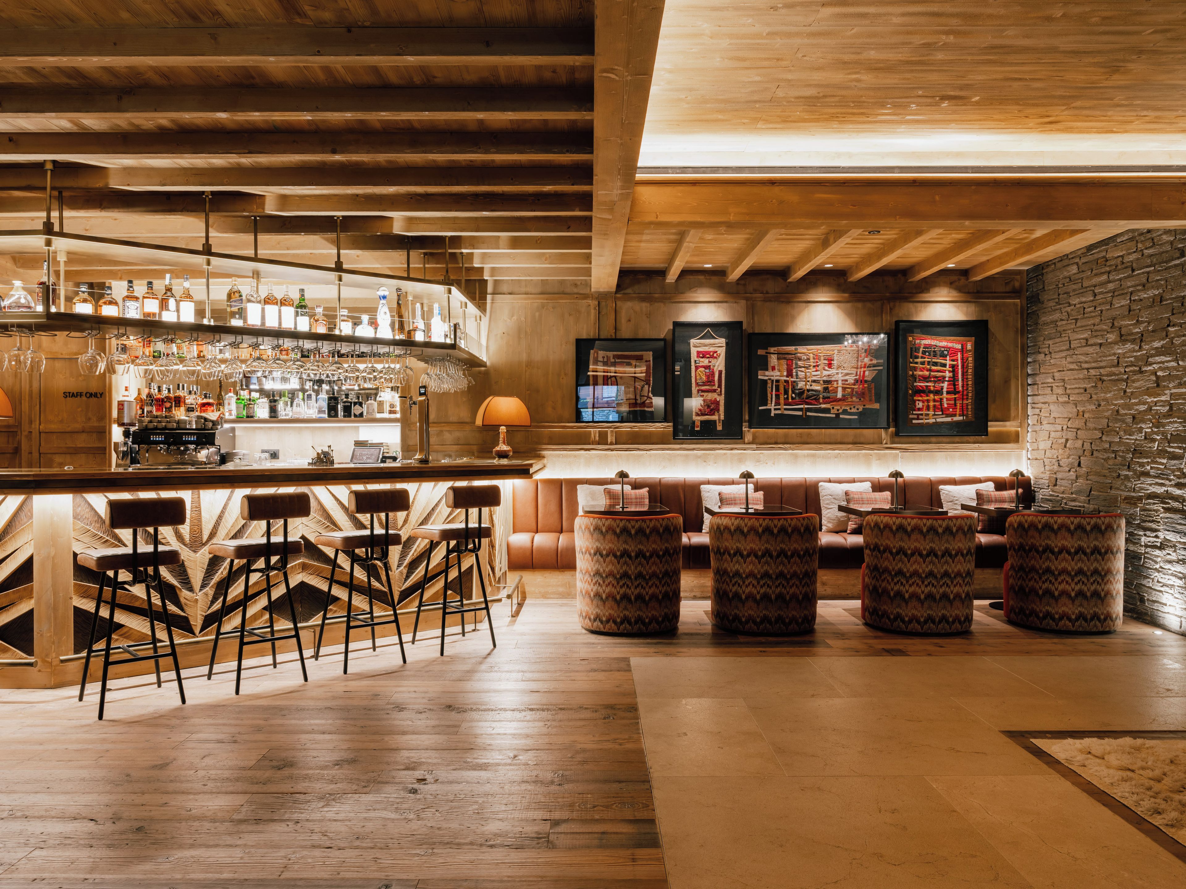 A bar area with wooden beams, a bar counter, stools, and a seating area with cushioned chairs.