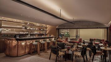 Un moderno bar con barra de cobre, taburetes de madera y una variedad de botellas y vasos. Hay varias zonas de estar con sillas acolchadas y mesas, y un amplio ventanal con vistas al exterior.