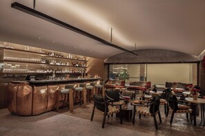 Un moderno bar con barra de cobre, taburetes de madera y una variedad de botellas y vasos. Hay varias zonas de estar con sillas acolchadas y mesas, y un amplio ventanal con vistas al exterior.
