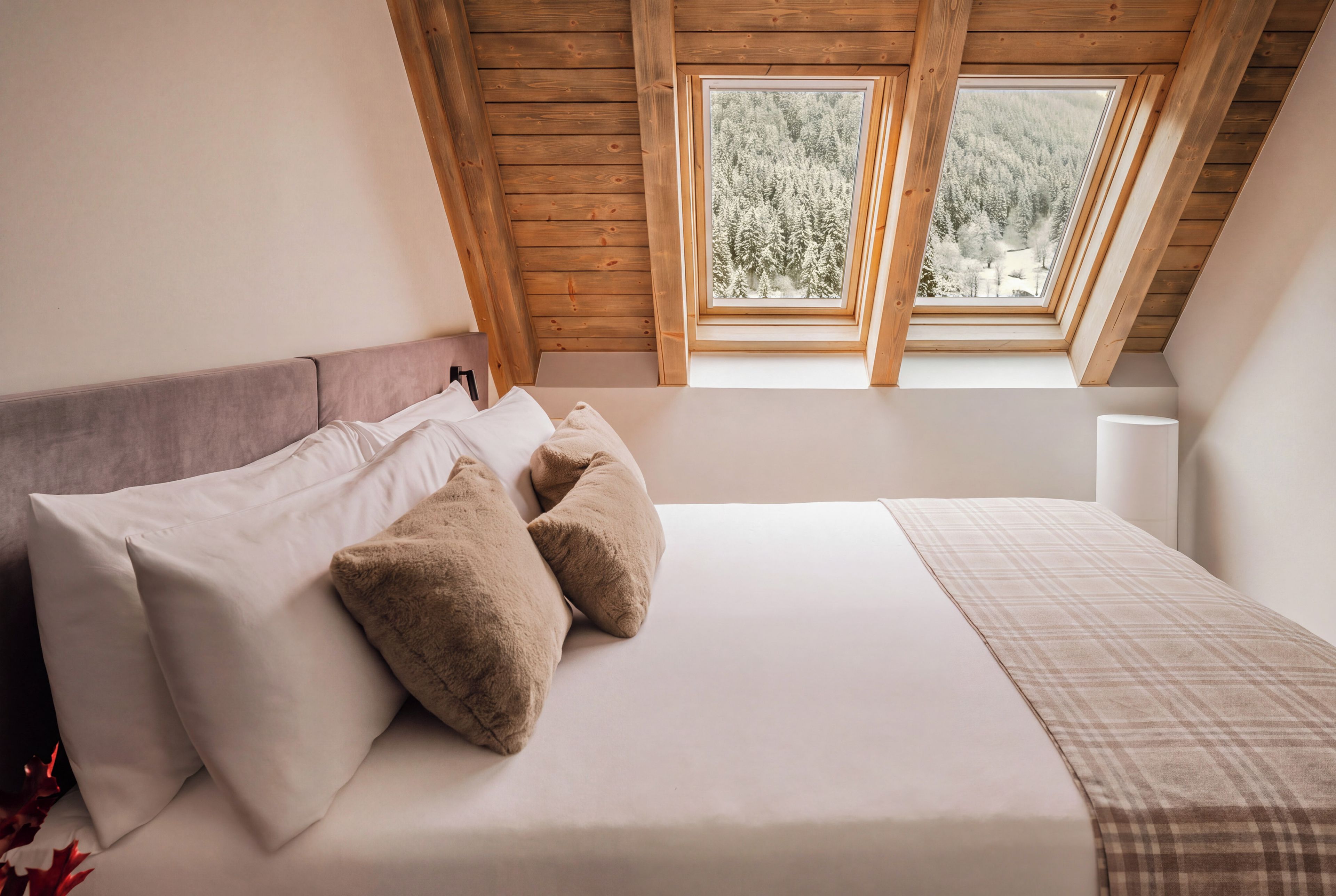 Ein Schlafzimmer mit schrägem Dach, einem Bett mit weißer Bettwäsche und zwei Kissen, und Blick auf einen Wald durch ein Dachfenster.