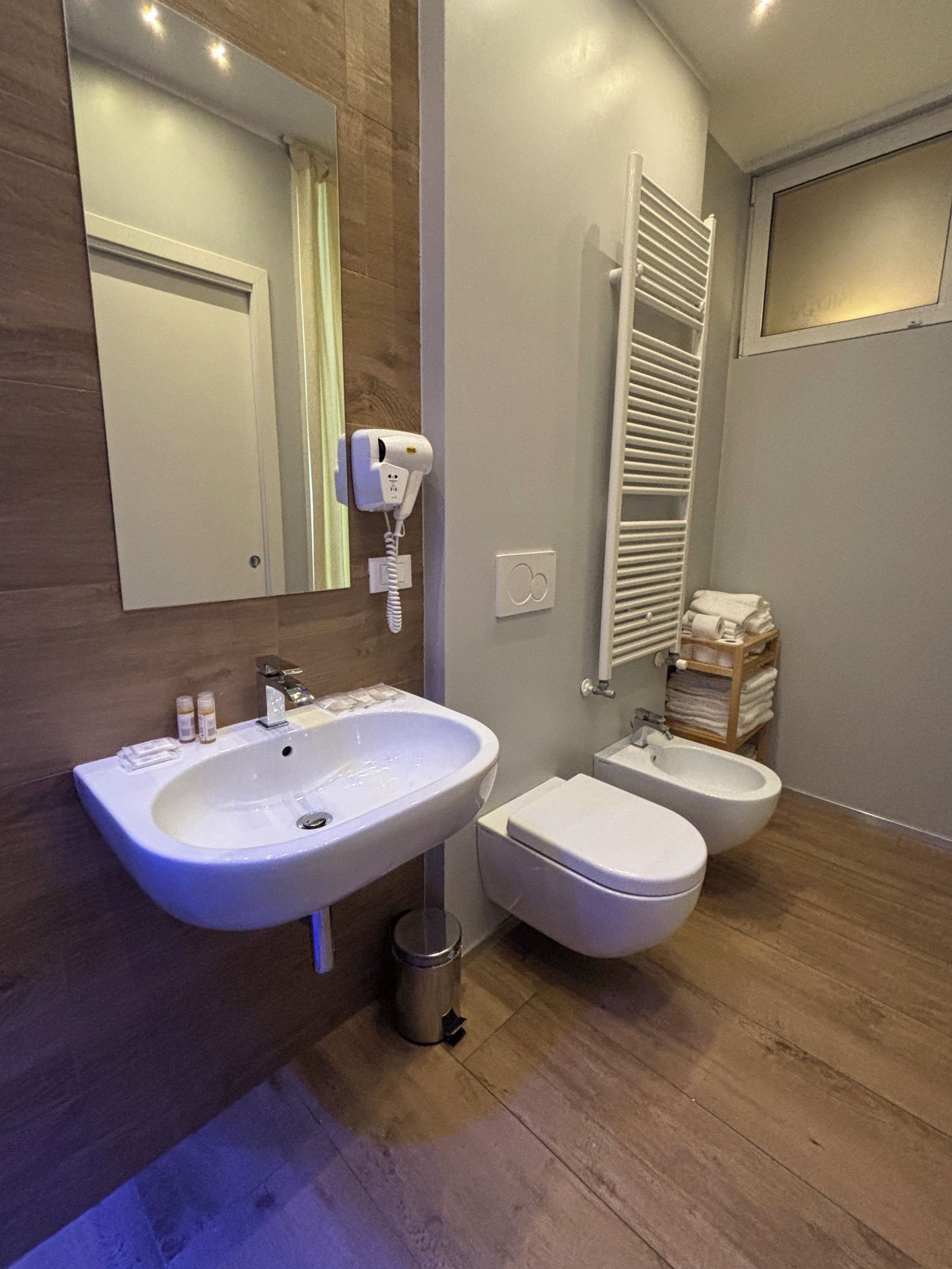 Une salle de bain moderne avec un grand lavabo, des toilettes et un bidet. Un miroir, un porte-serviettes et un sèche-cheveux sont fixés au mur.