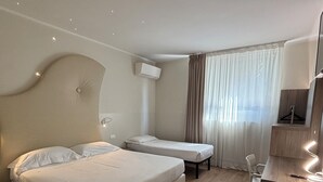 Quarto de hotel com duas camas, uma escrivaninha, uma cadeira, ar condicionado e uma janela com cortinas.
