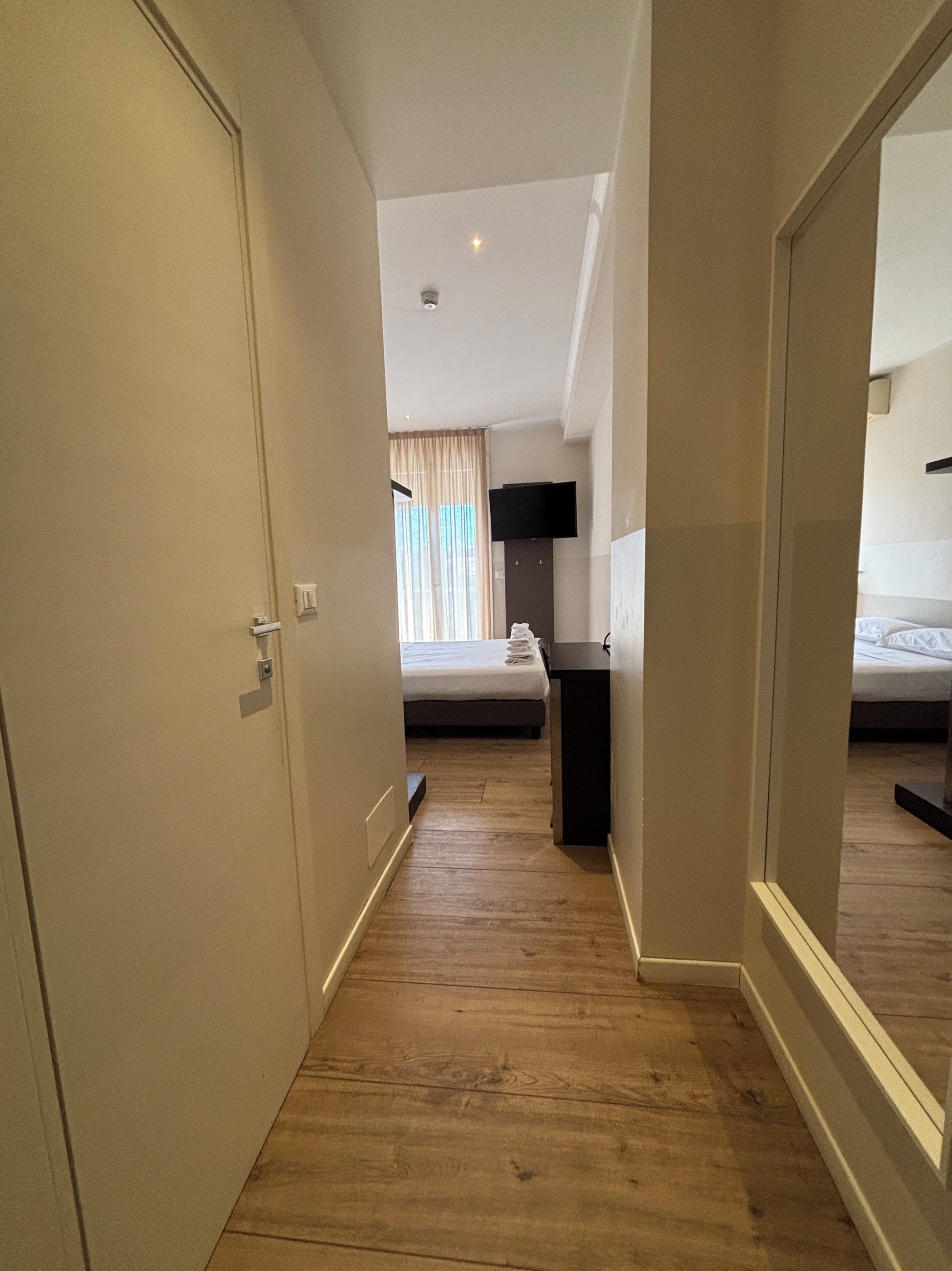 Un pasillo de hotel con piso de madera, una cama con colchón blanco, un televisor de pantalla plana montado en la pared y una puerta a la izquierda.