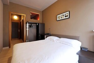 Habitación de hotel con cama, escritorio, refrigerador, espejo y una pequeña estantería con fotografías.