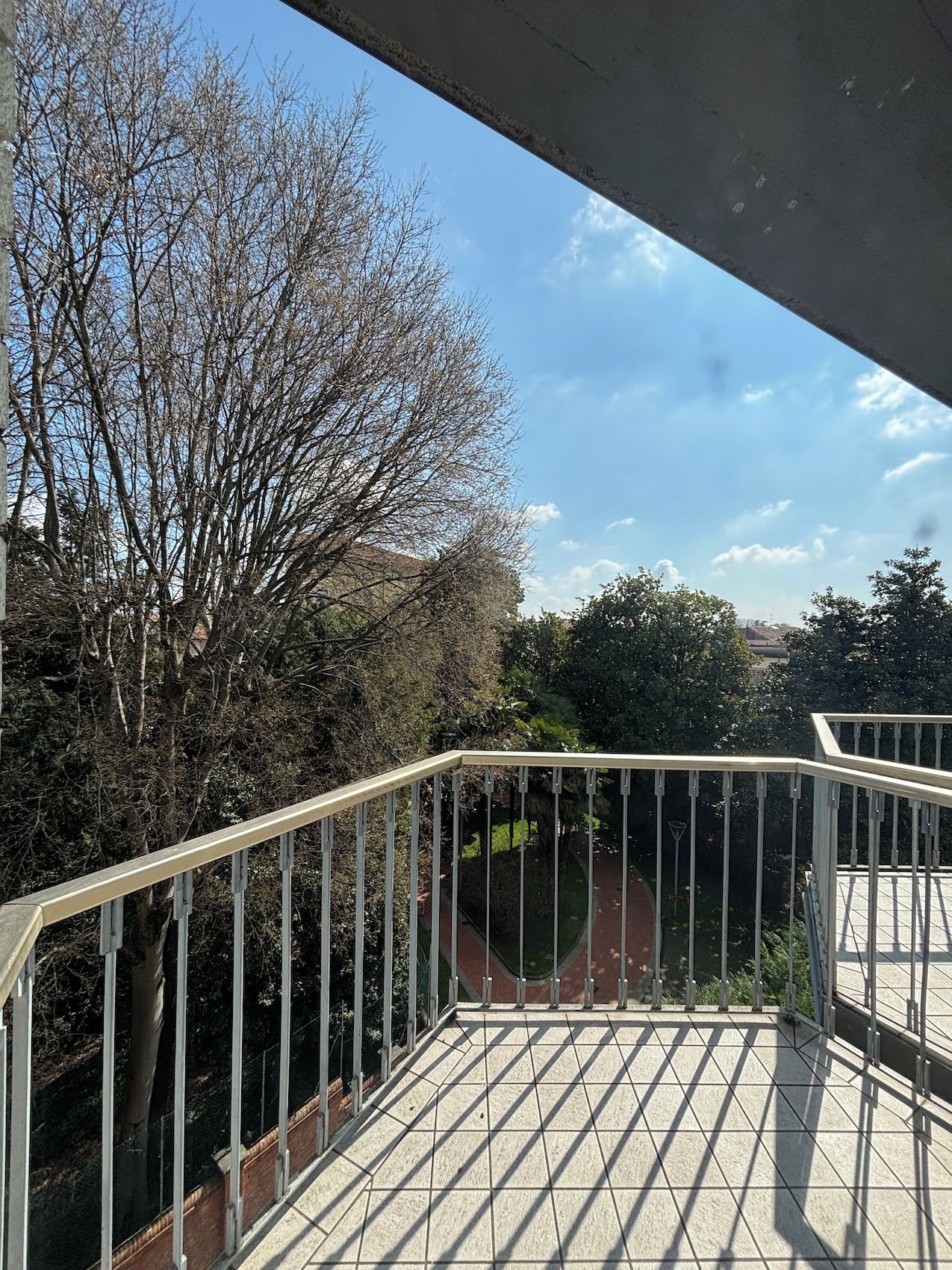 Un balcó amb paviment de rajoles i barana de metall, amb vista a un jardí i arbres.
