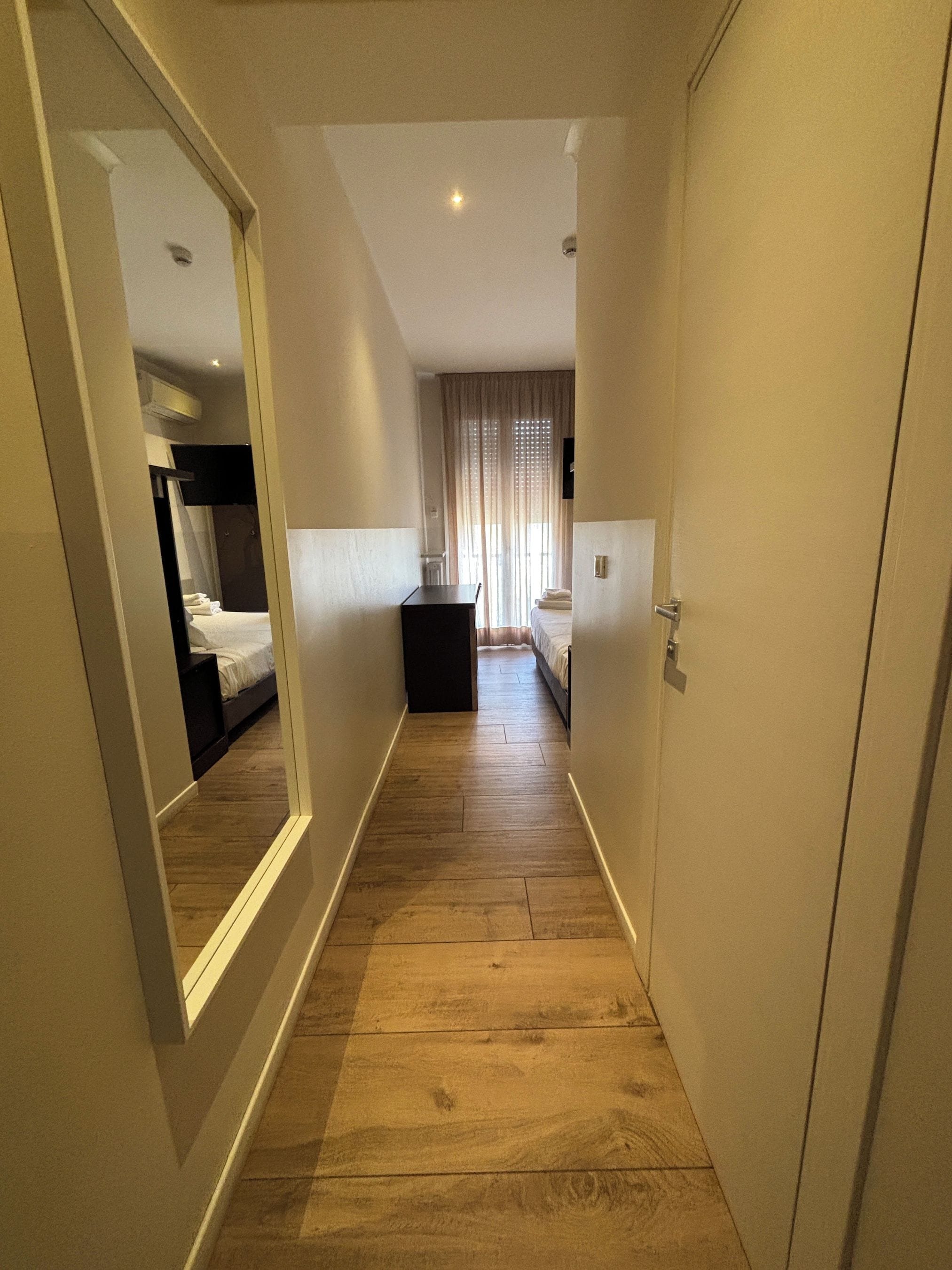 Pasillo de hotel con suelo de madera, una cama, un escritorio y un espejo que refleja la habitación.