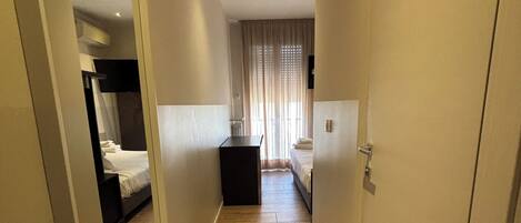 Pasillo de hotel con suelo de madera, una cama, un escritorio y un espejo que refleja la habitación.