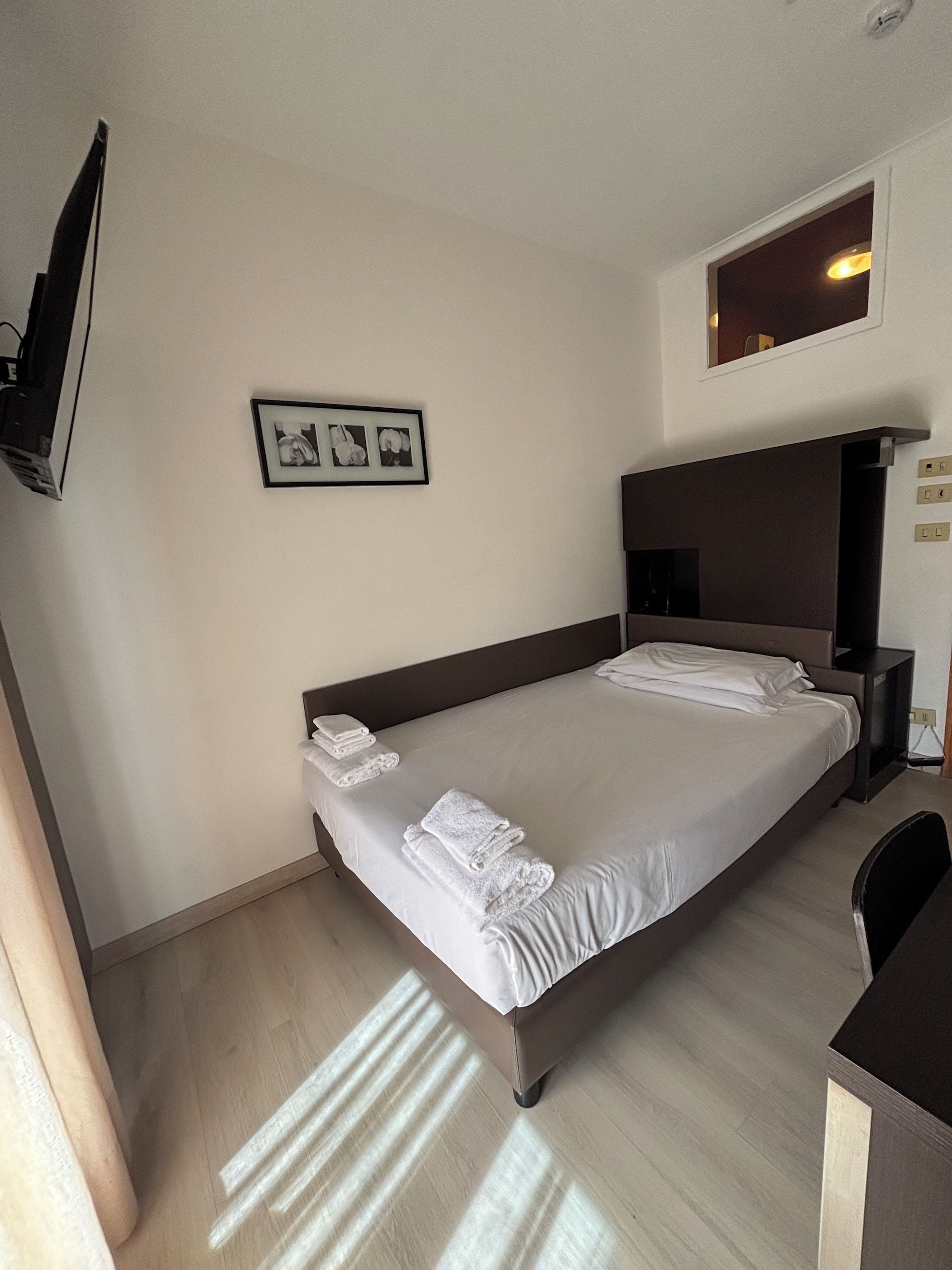 A hotel room with a bed, a flat-screen TV mounted on the wall, and a small window.