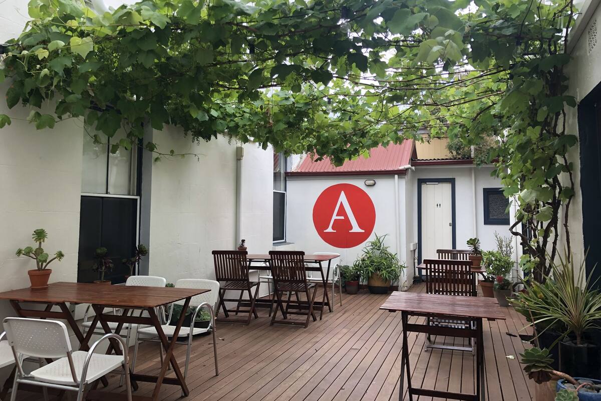 An outdoor patio area with wooden tables and chairs, surrounded by potted plants and greenery.
