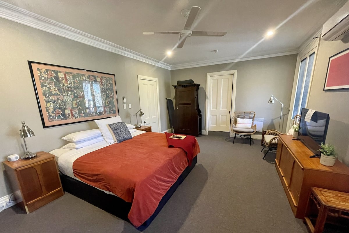 A bedroom with a bed, a ceiling fan, a desk, a chair, and a large framed picture on the wall.
