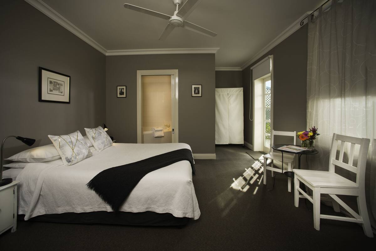 A hotel room with a large bed, a bedside table, a chair, a small table with a vase of flowers, and a door leading to a balcony.