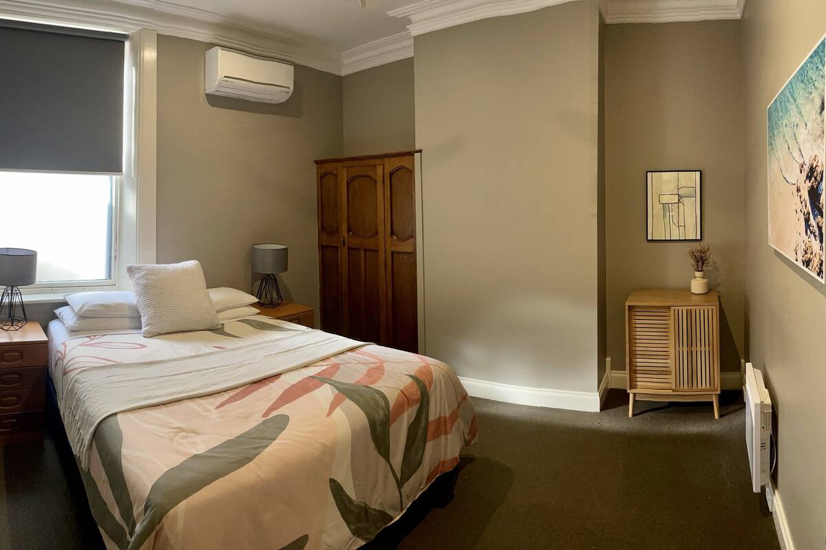 A bedroom with a bed, a nightstand, a dresser, an air conditioning unit, a radiator, and a painting on the wall.