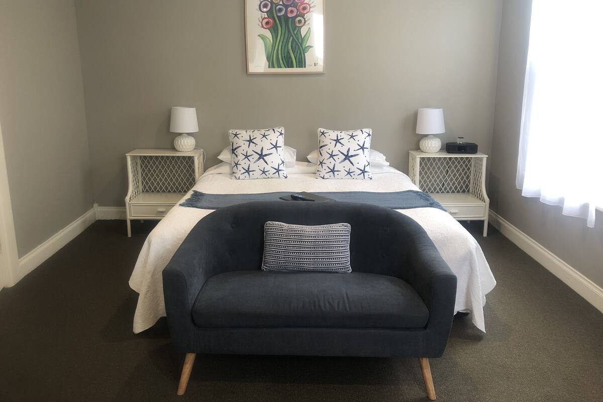 A bedroom with a bed, two bedside tables, a nightstand, a chair, and a wall-mounted picture.