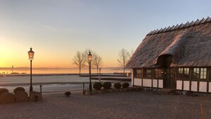 A thatched-roof building with a gravel pathway, a lamp post, and a view of a body of water at sunset. - Munkebo Kro (Munkebo)