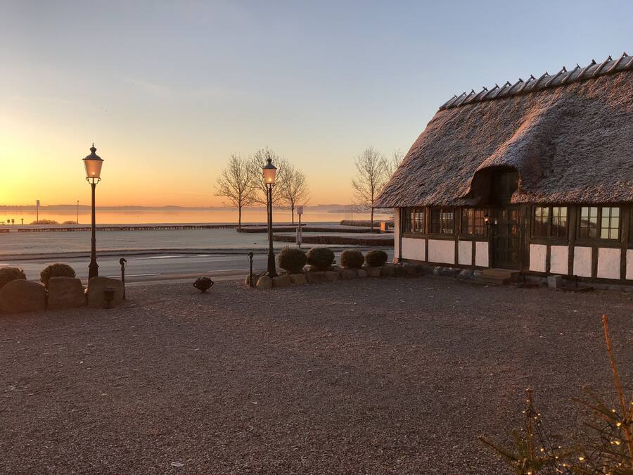 A thatched-roof building with a gravel pathway, a lamp post, and a view of a body of water at sunset.