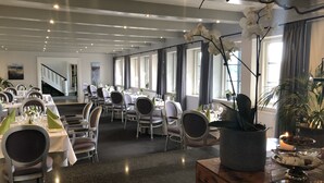 A spacious dining area with multiple tables set for guests, featuring a modern chandelier, large windows with curtains, and a staircase in the background. - Munkebo Kro (Munkebo)