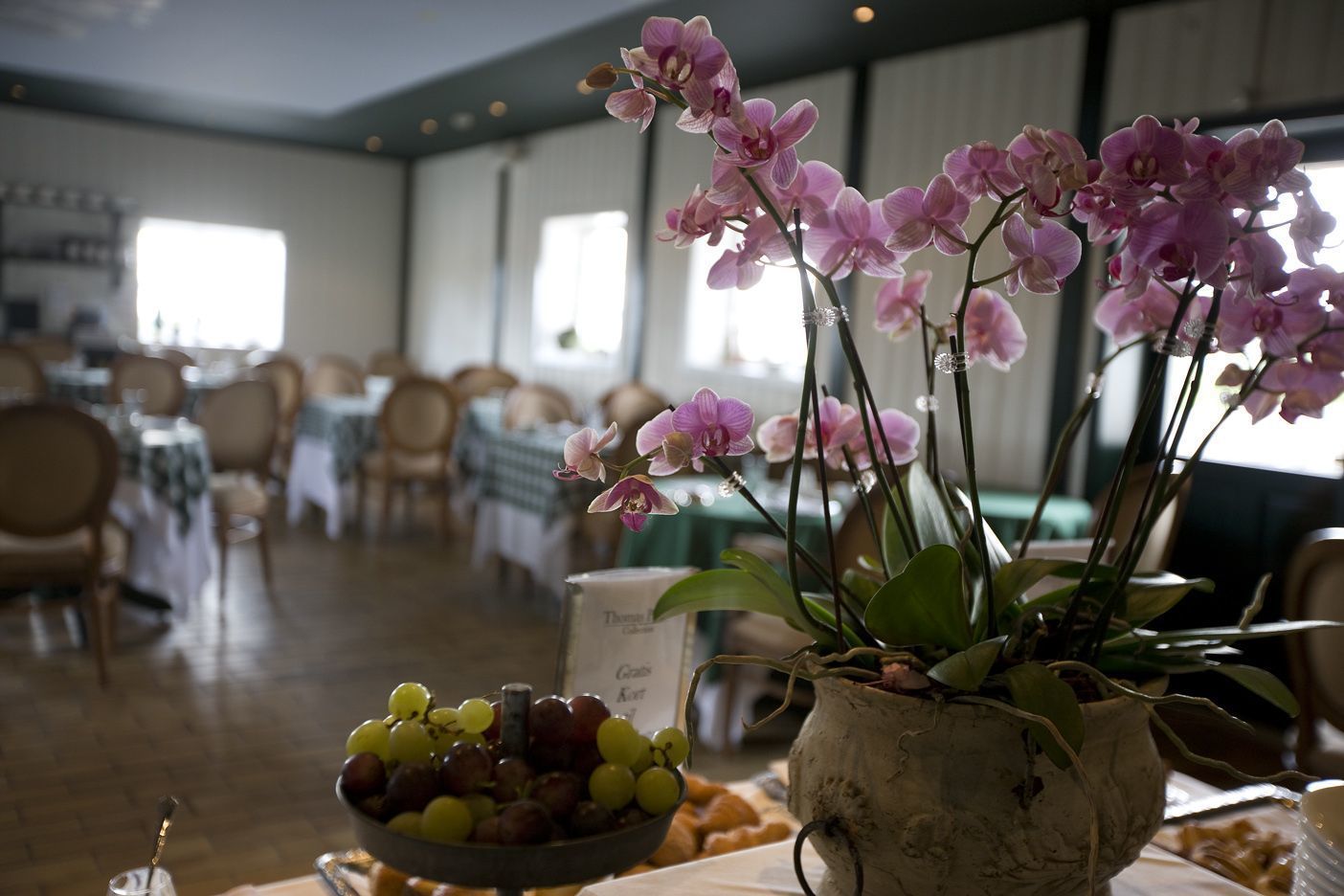 Un comedor con mesas dispuestas para los huéspedes, un cuenco de uvas y una orquídea en maceta.
