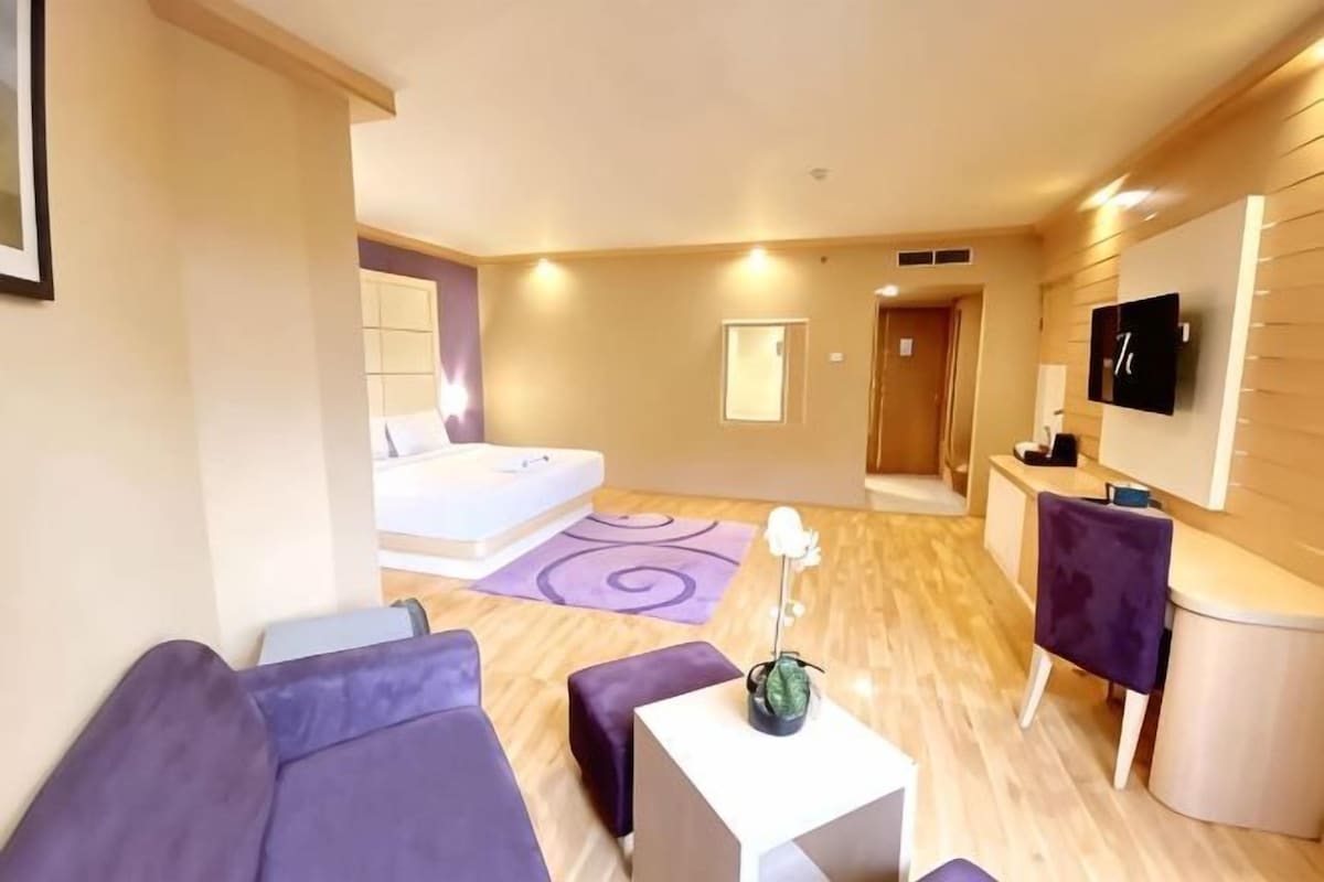 A modern hotel room with a bed, a seating area with a sofa and armchairs, a small table, and a TV mounted on the wall.