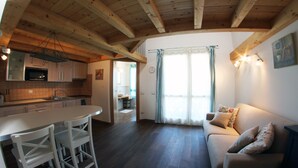 Et moderne køkken med træbjælker, et spiseområde med hvide stole, en sofa med dekorative puder og et stort vindue med gardiner.