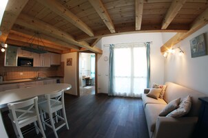 Una cucina moderna con travi in legno, un'area pranzo con sedie bianche, un divano con cuscini decorativi e un'ampia finestra con tende.
