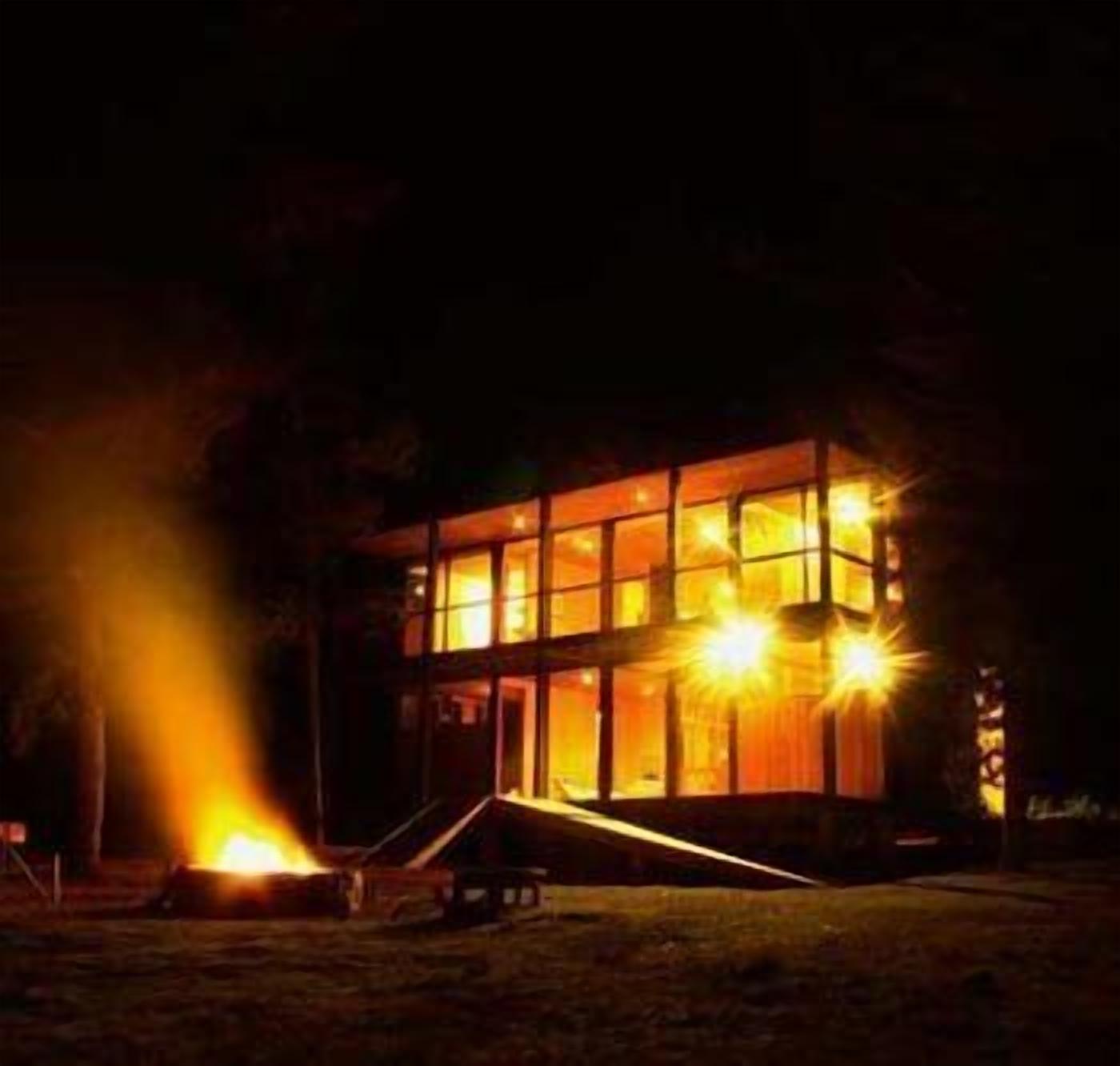 A modern building with large windows and a warm interior glow, surrounded by a grassy area with a fire.