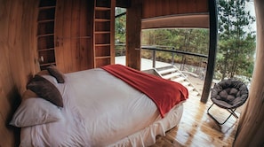 A wooden cabin bedroom with a bed, pillows, a red blanket, a chair, and a view of trees through a large window. - Surf Lodge Posada Punta de Lobos (Pichilemu)