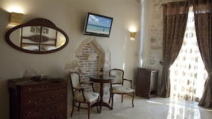 A room with a stone wall, a wooden dresser, two chairs, a small table, a television, and a window with curtains. - Antica Dimora Suites (Rethymno)