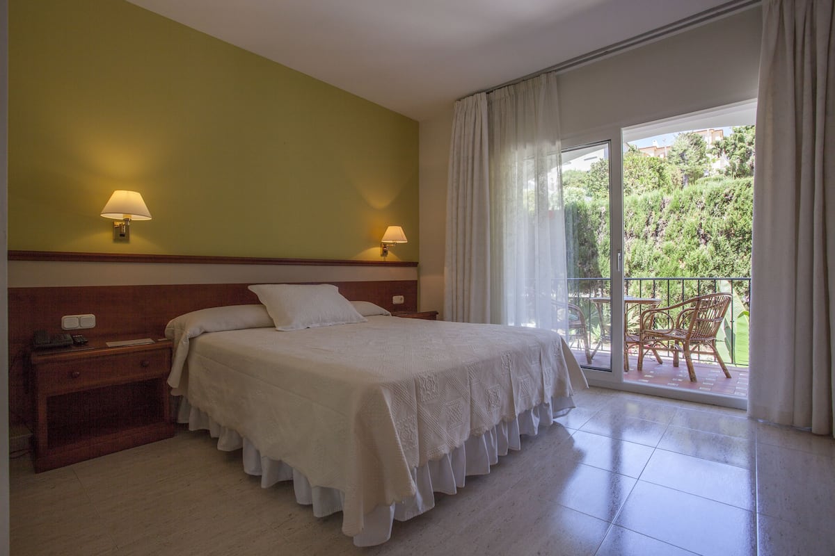 A hotel room with a bed, nightstand, and a view of a balcony with chairs.