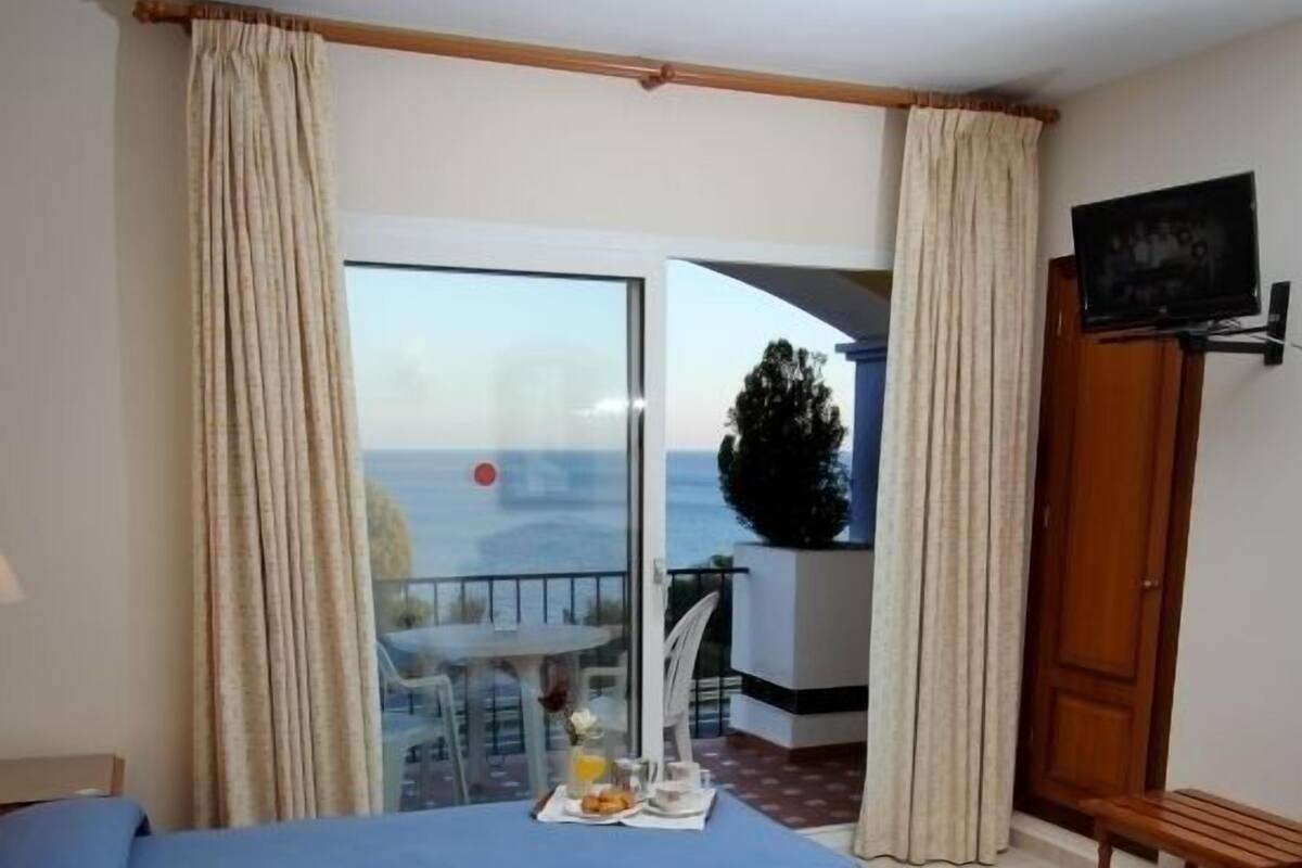 A hotel room with a large bed, a small table with a tray of food, a TV, and a balcony with a view of the sea.