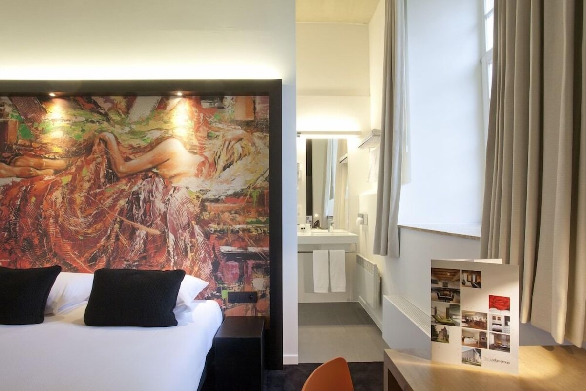Een hotelkamer met een groot abstract schilderij aan de muur, een bed met zwarte kussens, een kleine tafel met een brochure, en een badkamer die door een open deur zichtbaar is.