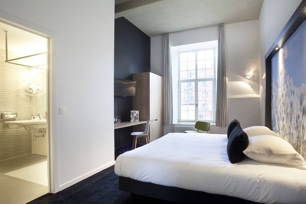 Une chambre d’hôtel moderne, dotée d’un grand lit, d’un bureau, d’une chaise, d’une petite table et d’une fenêtre avec des rideaux.
