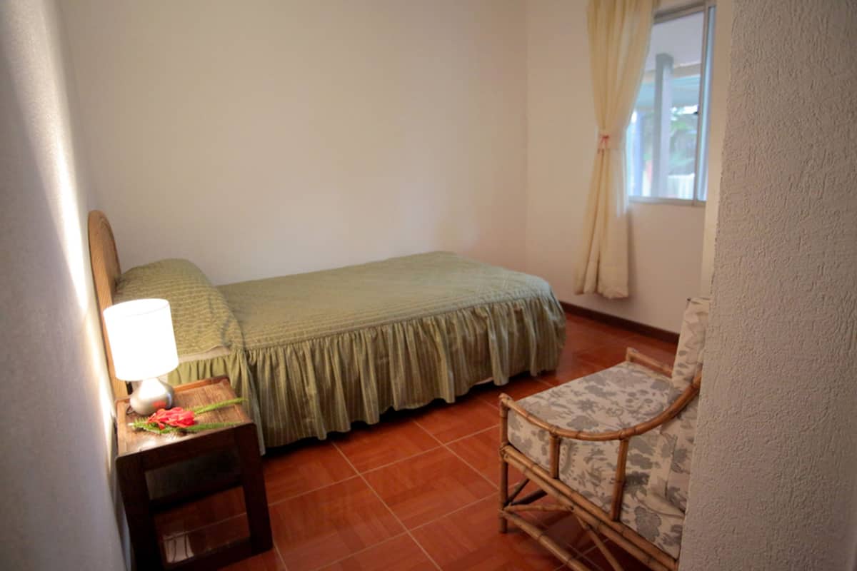 A small bedroom with a bed, a chair, a nightstand with a lamp, and a window with curtains.