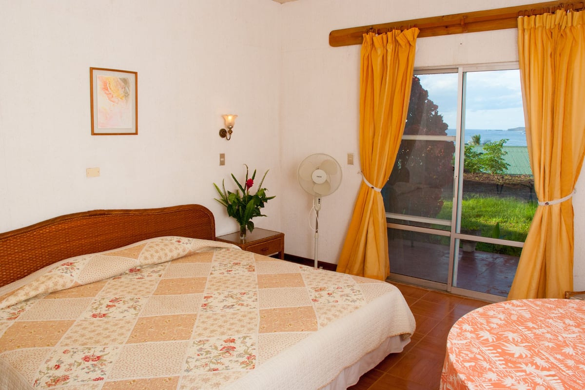 A bedroom with a bed, a bedside table with a lamp, a window with curtains, and a view of the outdoors.