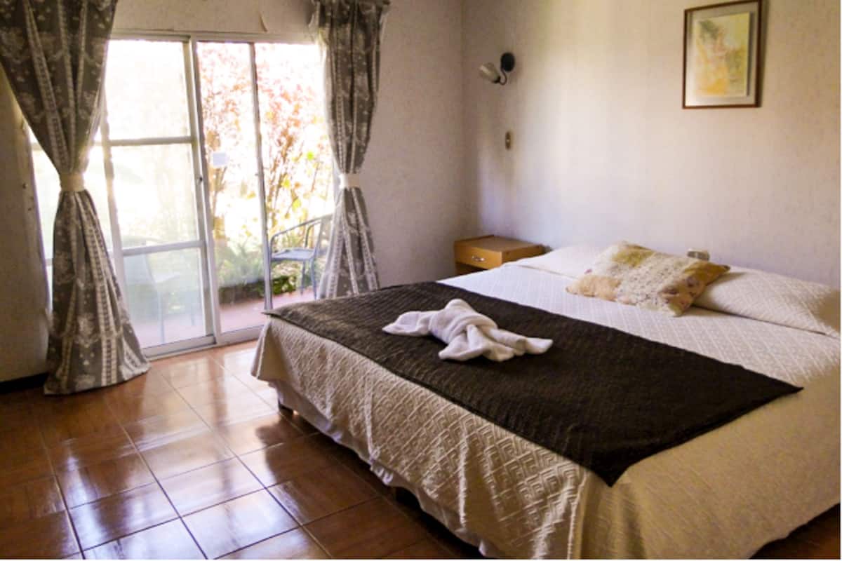 A bedroom with a bed, a nightstand, a window with curtains, and a door leading to an outdoor area.