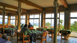 A restaurant with wooden interiors and large windows offering ocean views. Tables are set with chairs, and the space is well-lit. - Tupa Hotel (Hanga Roa)