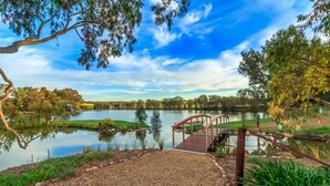 A gravel pathway leading to a wooden bridge over a calm lake, surrounded by trees and a clear sky.