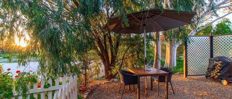 Een buitenzitgedeelte met een tafel en stoelen onder een pergola, omgeven door bomen en een witte hekken.